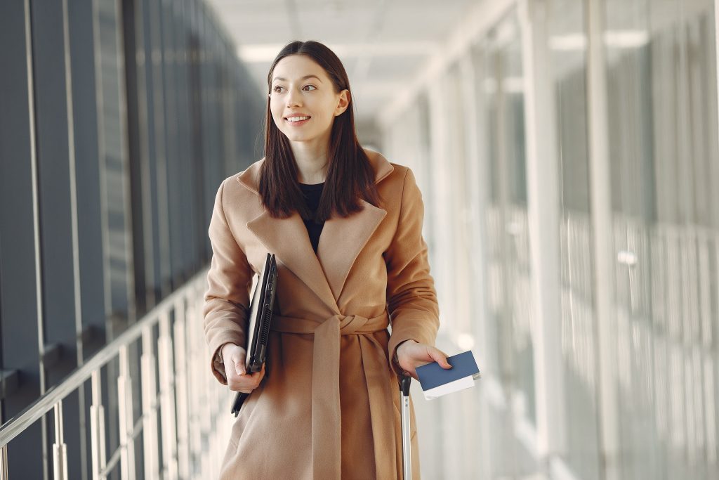  A imagem mostra uma mulher em um aeroporto segurando um passaporte e um notebook. 